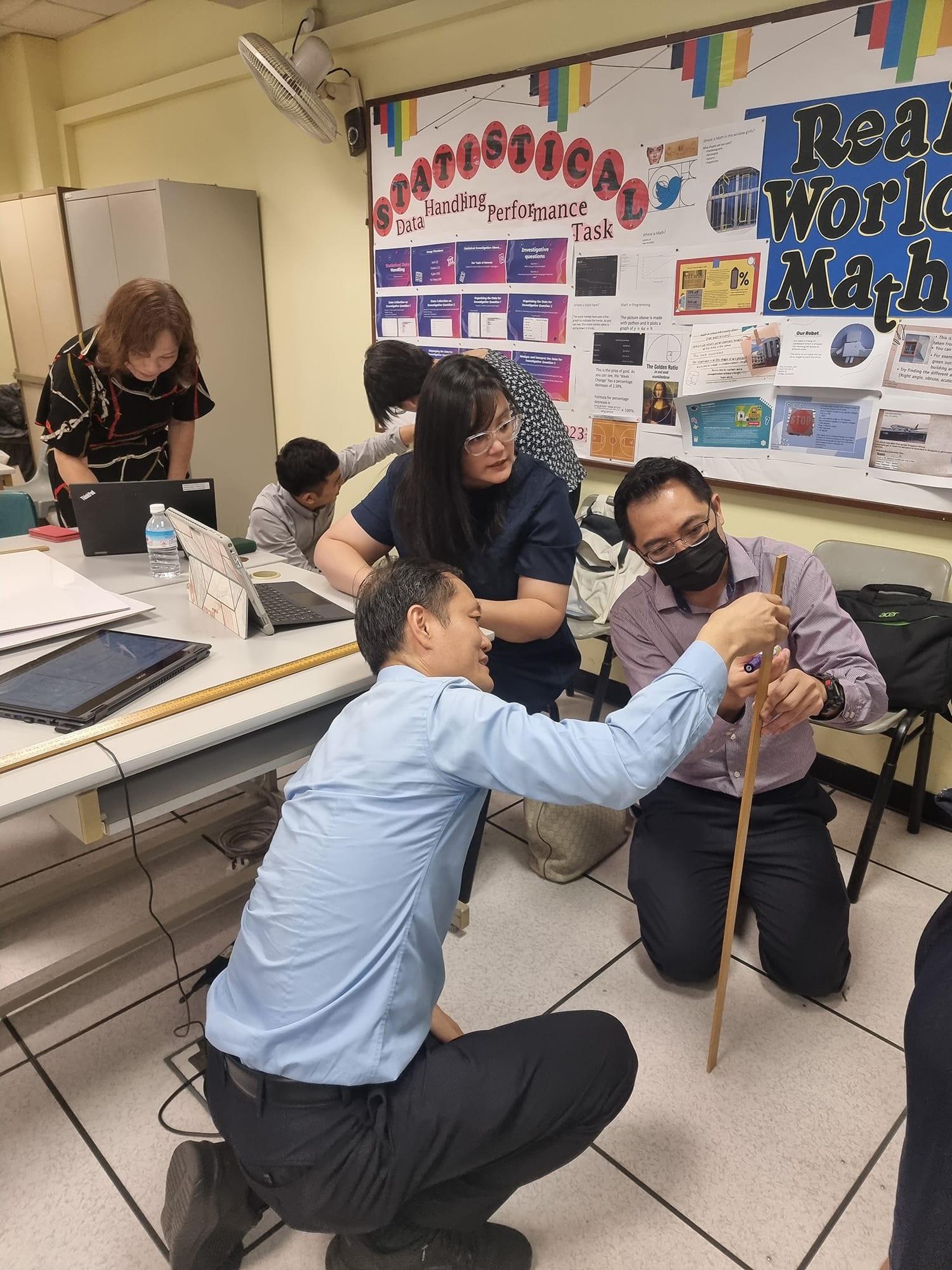 Three adults in a classroom examine a wooden stick near a bulletin board labeled "Statistical Data Handling Performance Task."